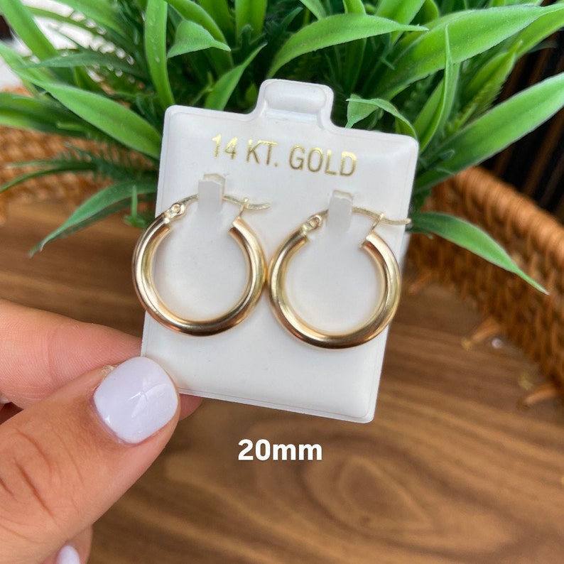 Puede incluir: Un par de pendientes de aro de oro de 14 quilates, de 20 mm de di&aacute;metro. Los pendientes se muestran en una tarjeta blanca con el texto "14 KT. GOLD" y "20mm".