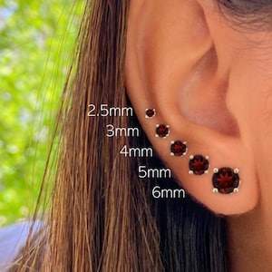 May include: A close-up of a person's ear with five red gemstone earrings of different sizes. The earrings are labeled with their sizes in millimeters: 2.5mm, 3mm, 4mm, 5mm, and 6mm.