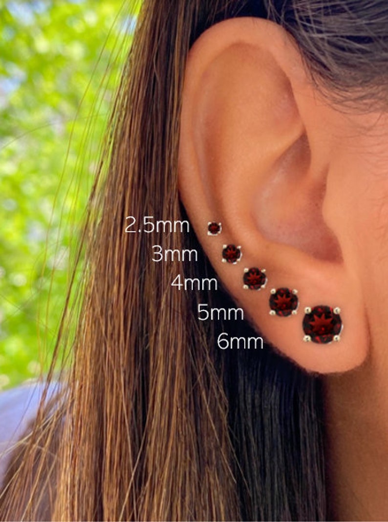 May include: A close-up of a person's ear with five red gemstone earrings of different sizes. The earrings are labeled with their sizes in millimeters: 2.5mm, 3mm, 4mm, 5mm, and 6mm.
