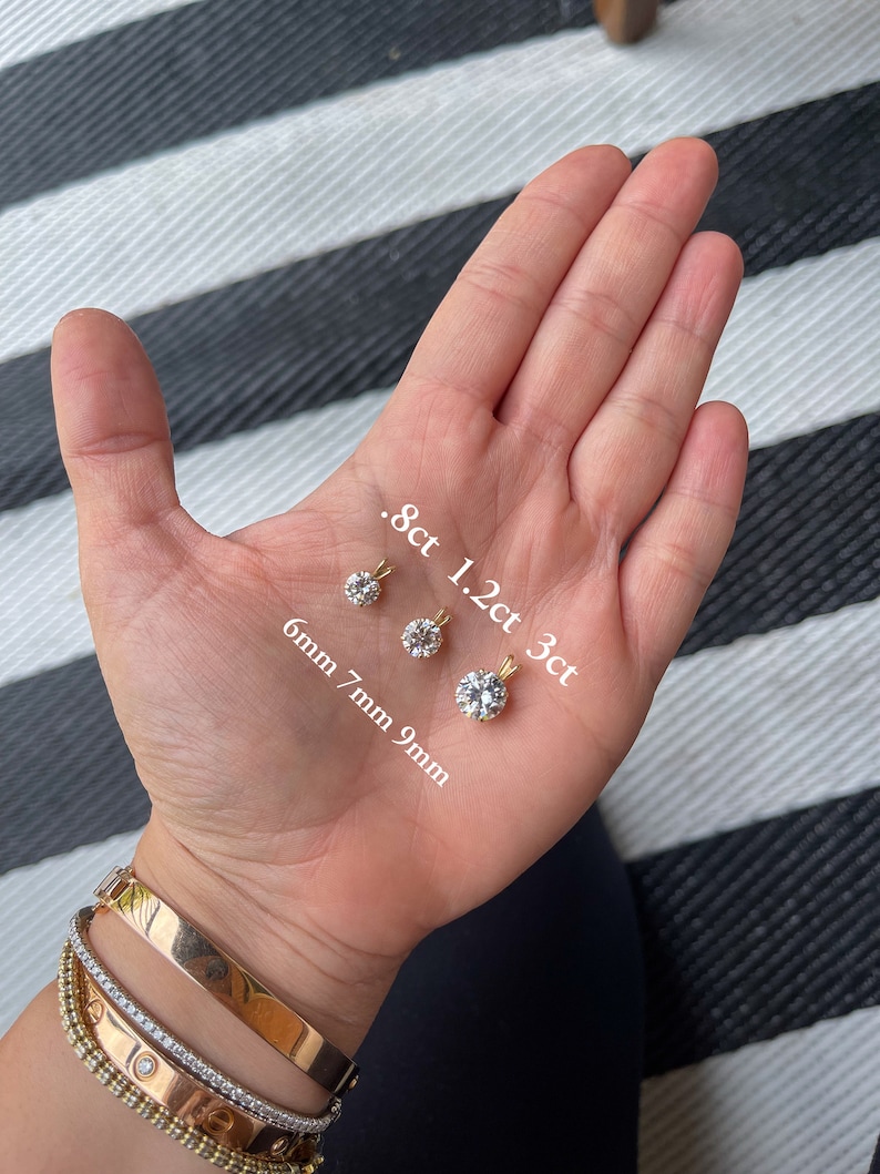 May include: Three gold pendants with different sized round diamonds. The pendants are labeled with their carat weight: .8ct, 1.2ct, and 3ct. The pendants are also labeled with their size in millimeters: 6mm, 7mm, and 9mm.