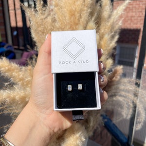 May include: A white box with the text "ROCK A STUD" on the top. The box contains a pair of small, rectangular, clear earrings.
