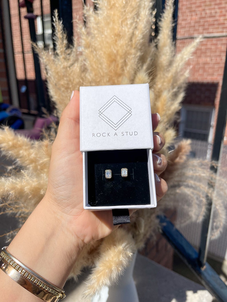 May include: A white box with the text "ROCK A STUD" on the top. The box contains a pair of small, rectangular, clear earrings.
