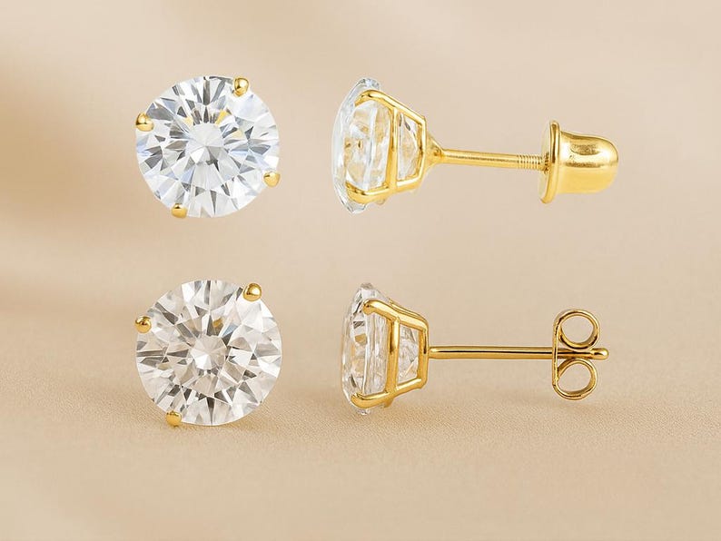 May include: A pair of round, clear gemstone stud earrings with gold-colored settings and posts. The earrings are displayed in two different orientations, showcasing the sparkling facets of the gemstones and the details of the gold backing.