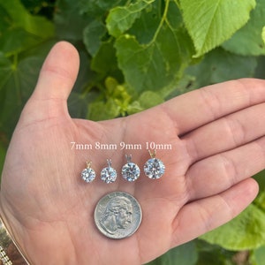 May include: Four round, clear, cubic zirconia pendants in different sizes, 7mm, 8mm, 9mm, and 10mm, on a hand with a US quarter for size comparison.