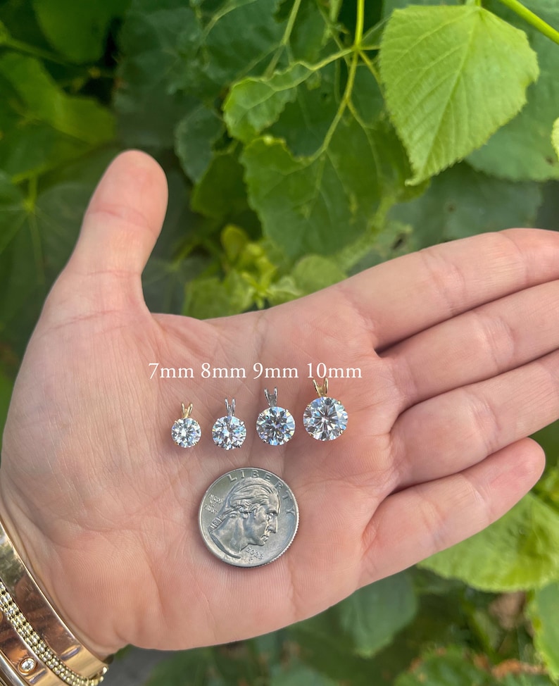 May include: Four round, clear, cubic zirconia pendants in different sizes, 7mm, 8mm, 9mm, and 10mm, on a hand with a US quarter for size comparison.