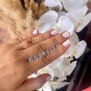 May include: A collection of round, clear gemstones of varying sizes, arranged in a line. The gemstones are displayed against a background of white flowers and a brick wall. The gemstones are sparkling and reflective.