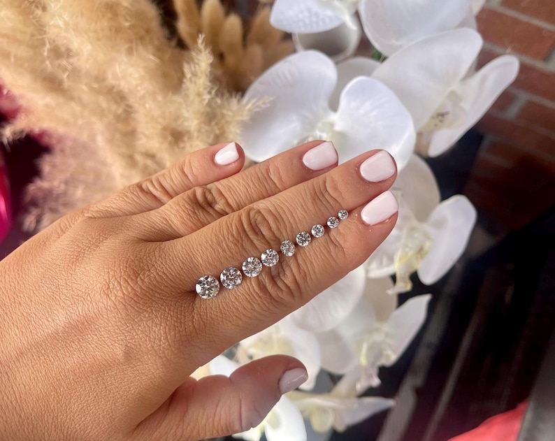 May include: A collection of round, clear gemstones of varying sizes, arranged in a line. The gemstones are displayed against a background of white flowers and a brick wall. The gemstones are sparkling and reflective.