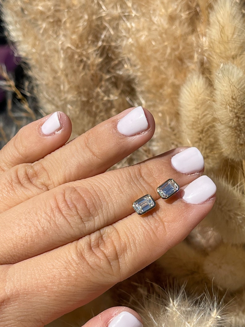 May include: A pair of emerald cut diamond earrings in a gold setting. The earrings are shown on a hand with white nail polish.