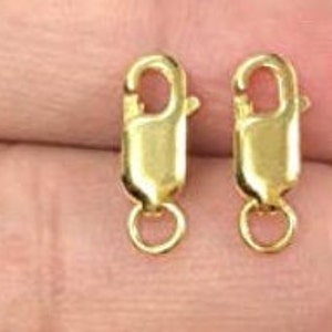 K&ouml;nnte beinhalten: Zwei goldfarbene Metall-Hummer-Verschl&uuml;sse mit einem runden Ring an jeder Seite. Die Verschl&uuml;sse werden h&auml;ufig f&uuml;r die Schmuckherstellung verwendet.