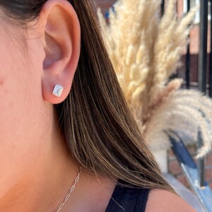 May include: A close-up of a person's ear with a small, clear, emerald-cut gemstone earring. The earring is set in a gold setting.