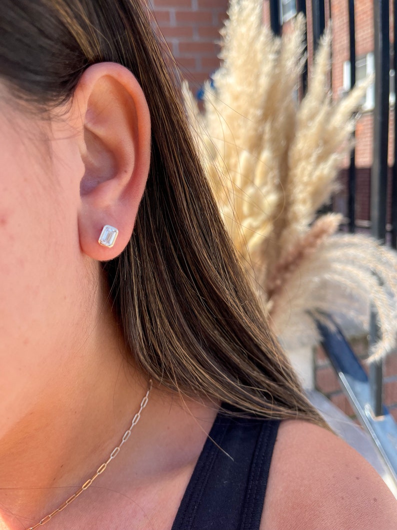 May include: A close-up of a person's ear with a small, clear, emerald-cut gemstone earring. The earring is set in a gold setting.