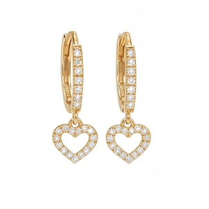 May include: A pair of gold-colored hoop earrings. Each earring features a heart-shaped charm, outlined with small, clear gemstones. The hoops are also embellished with similar gemstones. The earrings are displayed against a white background.