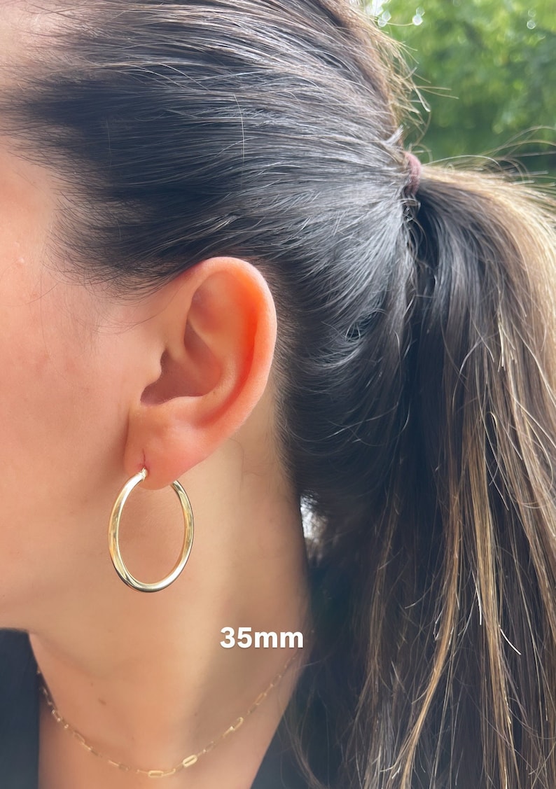 Puede incluir: Pendientes de aro de oro de 35 mm de di&aacute;metro. Los pendientes se muestran en la oreja de una persona.