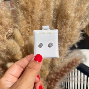 May include: A pair of round-cut diamond stud earrings on a white display card. The card is labeled "14 KT. GOLD". The earrings are displayed against a backdrop of dried, tan-colored ornamental grass.
