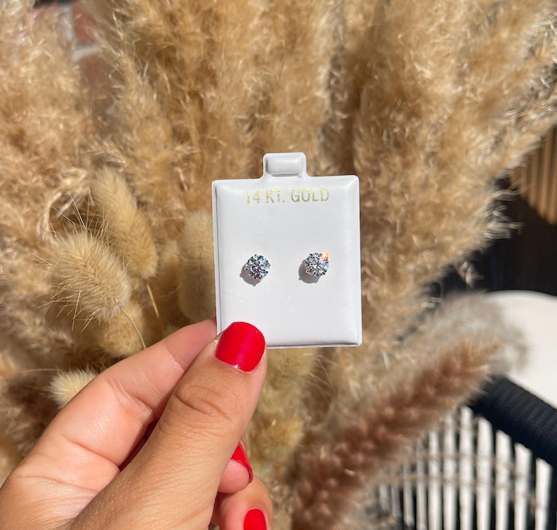 May include: A pair of round-cut diamond stud earrings on a white display card. The card is labeled "14 KT. GOLD". The earrings are displayed against a backdrop of dried, tan-colored ornamental grass.