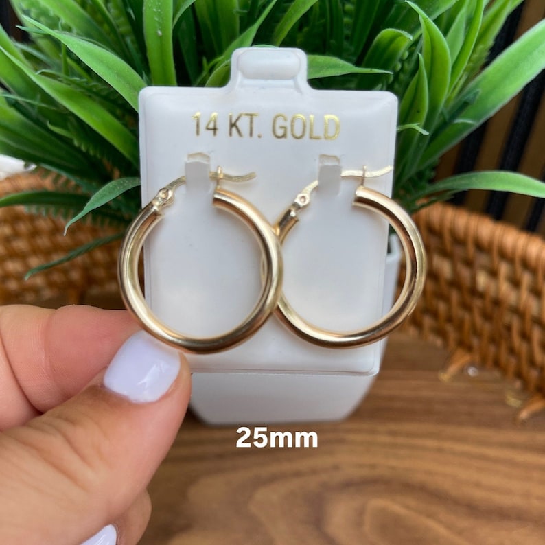 Puede incluir: Un par de pendientes de aro de oro de 14 quilates con un di&aacute;metro de 25 mm. Los pendientes est&aacute;n en una tarjeta blanca con el texto "14 KT. GOLD" y "25mm".