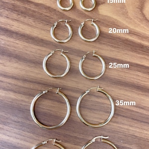 Puede incluir: Cinco pares de pendientes de aro de oro de diferentes tama&ntilde;os, que van desde 15 mm hasta 40 mm de di&aacute;metro. Los pendientes se muestran sobre una superficie de madera.