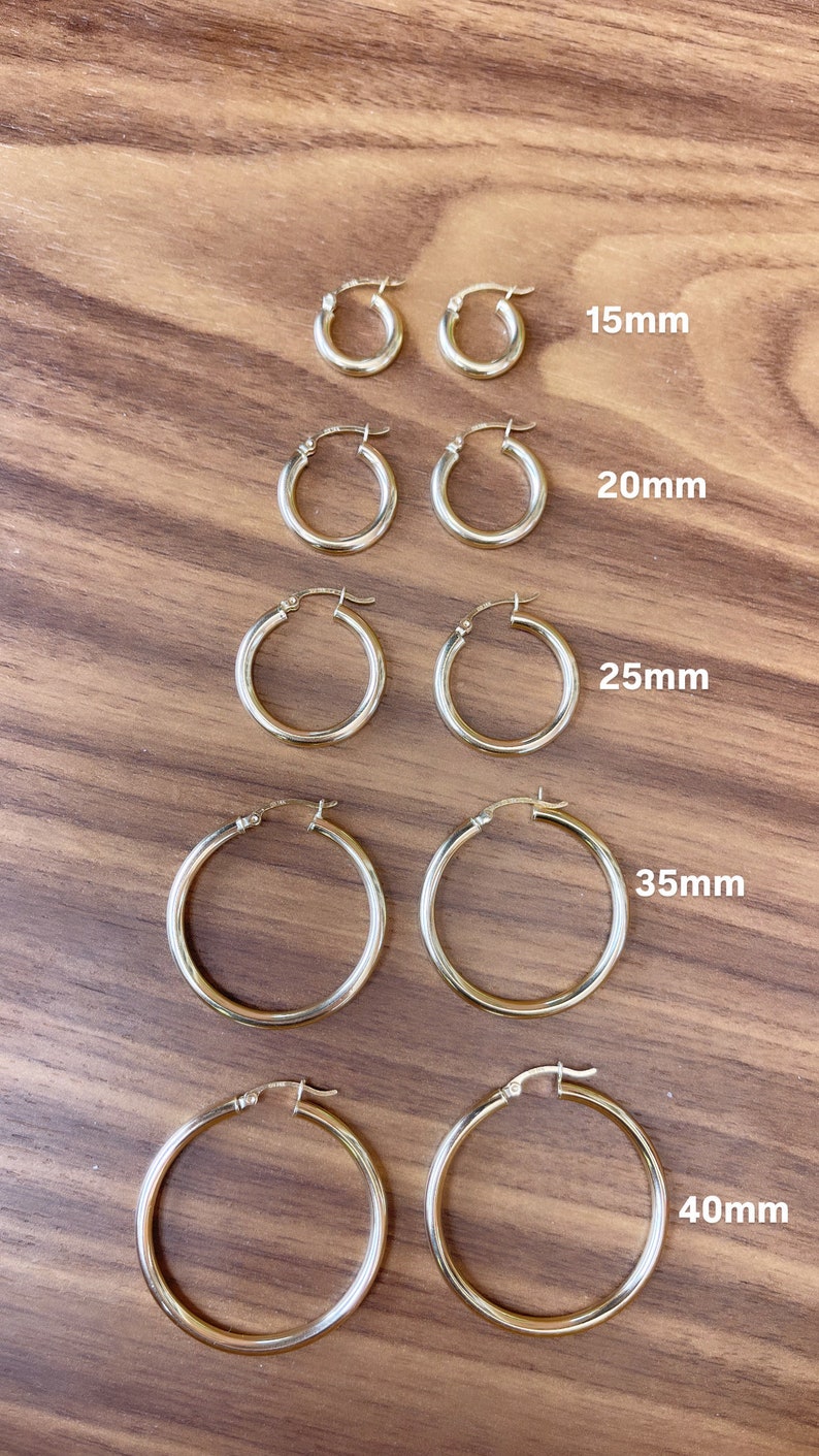Puede incluir: Cinco pares de pendientes de aro de oro de diferentes tama&ntilde;os, que van desde 15 mm hasta 40 mm de di&aacute;metro. Los pendientes se muestran sobre una superficie de madera.