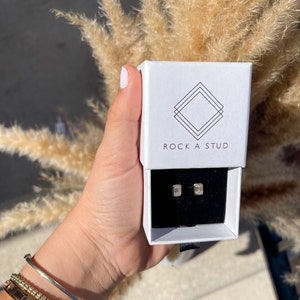 May include: A white box with the text "ROCK A STUD" on the front. The box contains a pair of small, rectangular, clear earrings.