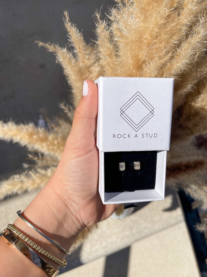 May include: A white box with the text "ROCK A STUD" on the front. The box contains a pair of small, rectangular, clear earrings.