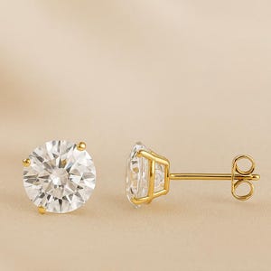 May include: A pair of gold stud earrings with large, round, clear gemstones. The gemstones are faceted, reflecting light. The earrings have a classic design with a post and butterfly backing.