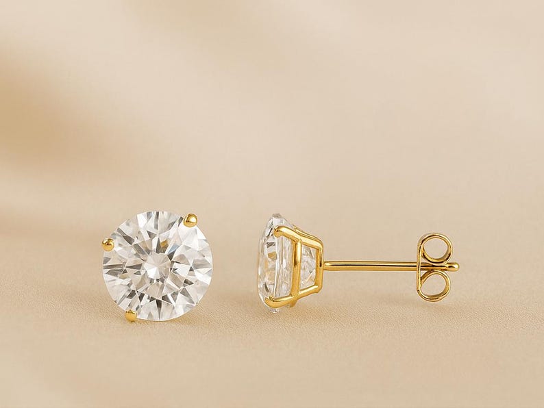 May include: A pair of gold stud earrings with large, round, clear gemstones. The gemstones are faceted, reflecting light. The earrings have a classic design with a post and butterfly backing.