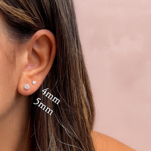 May include: Close-up of a person's ear with two small, round, diamond-like stud earrings. The earrings are 4mm and 5mm in size. The background is a soft pink color, and the person has long, brown hair.