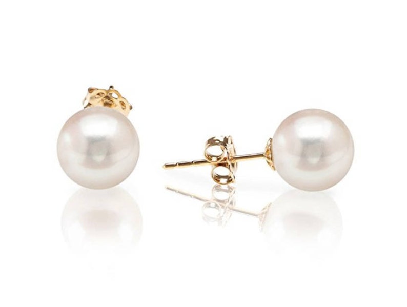 May include: A pair of stud earrings featuring large, round, white pearls set on gold posts. The earrings are displayed on a reflective white surface, showcasing their elegant design and luster. The gold posts have a decorative design.