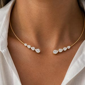 Puede incluir: Un collar dorado con un diseño delicado. El collar presenta una serie de piedras preciosas claras y brillantes, incluyendo piedras en forma de pera y redondas. El collar se lleva sobre una camisa blanca.