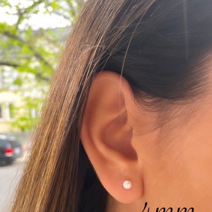 May include: A close-up of a person's ear with a small, round, white pearl stud earring. The earring is approximately 4mm in size. The person has long, brown hair. The background is blurred with green foliage.