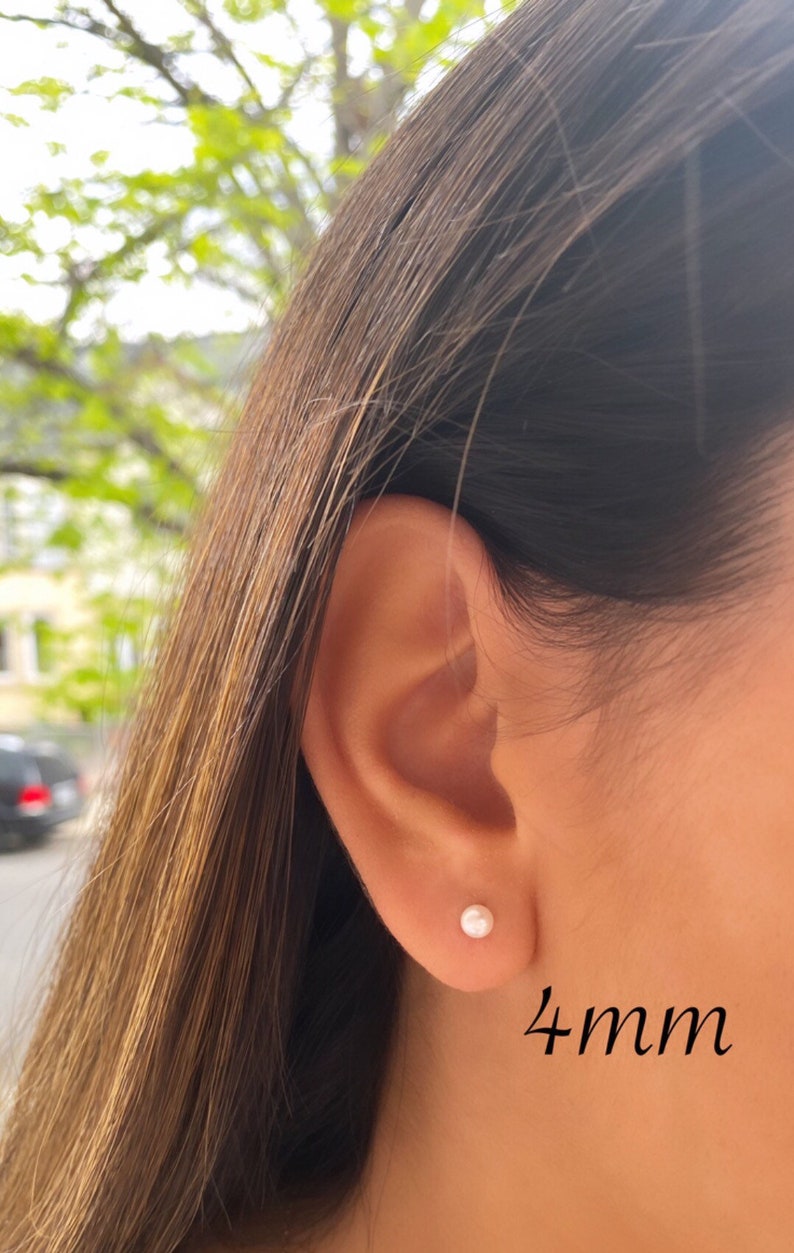 May include: A close-up of a person's ear with a small, round, white pearl stud earring. The earring is approximately 4mm in size. The person has long, brown hair. The background is blurred with green foliage.