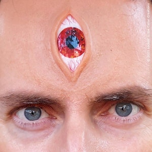 May include: A close-up of a person's face with a third eye painted on their forehead. The eye is red with a black pupil and white sclera. The person's other two eyes are blue.