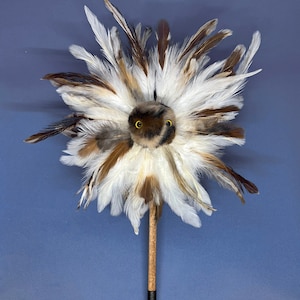 May include: A brown and white feather duster with a black handle and a small brown and white owl face with yellow eyes in the center of the duster.