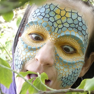 Könnte beinhalten: Eine Person mit aufwendiger Gesichtsbemalung, die ein blau-gelbes Reptilienschuppenmuster auf Stirn, Schläfen und um die Augen zeigt. Das Design verwendet texturierte, erhaben wirkende sechseckige Formen in Hellblau und Gold.