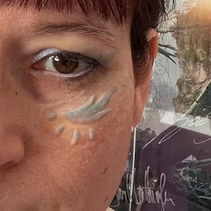 Puede incluir: Primer plano de un ojo con sombra de ojos plateada y un diseño de ala azul claro. La piel de la persona tiene un diseño de pluma y lágrima azul claro en la mejilla. El fondo está borroso.