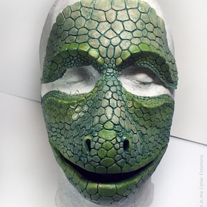 Op de afbeelding: Een groen reptielmasker met een gestructureerd, schubachtig patroon dat het voorhoofd, de neus en de mond bedekt. Het masker heeft uitsparingen voor de ogen en een kleine opening voor de mond. De tekst 2020 Locked In the Cellar Creations is zichtbaar.