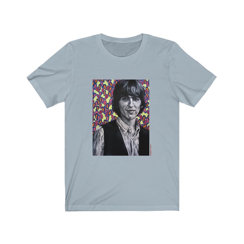 Puede incluir: Una camiseta azul claro con un estampado gr&aacute;fico de un hombre con cabello oscuro y un chaleco negro. El fondo del gr&aacute;fico es un patr&oacute;n colorido de rosa, amarillo y azul.