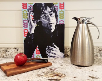 May include: A stainless steel thermal carafe with a silver handle and lid sits next to a canvas print of a black and white portrait of a man wearing glasses. A red apple and a wooden cutting board with a knife are in the foreground.