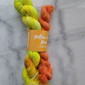 May include: A skein of hand-dyed yarn in vibrant yellow and orange hues. The yarn is bundled with a label that reads "Yellow Door Yarn Company". The yarn is displayed against a light gray marble background, showcasing its colors and texture.
