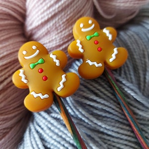 May include: Two gingerbread man stitch markers with green bow ties and red buttons, resting on colourful knitting needles. The markers are light brown with white icing details. The background is soft, textured yarn in shades of pink and grey.