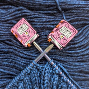 May include: Two pink enamel pins shaped like books with the text "BOOKISH GIRLIE" are crossed over each other. The pins are on knitting needles, with a partially knitted blue fabric in the foreground and blue yarn in the background.