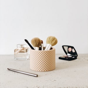 Puede incluir: Colección de productos de belleza sobre una superficie clara. Un recipiente cilíndrico texturizado de color melocotón contiene brochas de maquillaje. Un frasco de perfume con una cinta, una paleta de maquillaje y un lápiz plateado también están presentes.