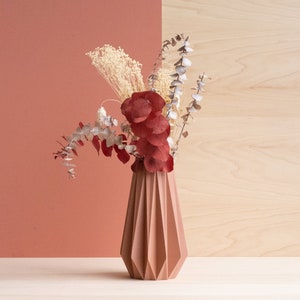 Peut inclure: Un vase en origami de couleur terre cuite avec un design géométrique contient un bouquet de fleurs séchées, dont de l'eucalyptus et des fleurs rouges.
