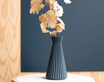 May include: A dark blue, pleated ceramic vase with a dried oak leaf arrangement on a white table with a wood base.