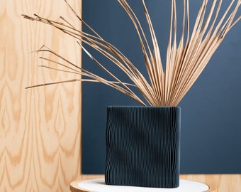 May include: A dark teal, square vase with a textured, wavy design, holding dried, tan-colored palm fronds. The vase sits on a small, round, white-topped table with a wooden base. The background features a wooden wall and a dark blue panel.