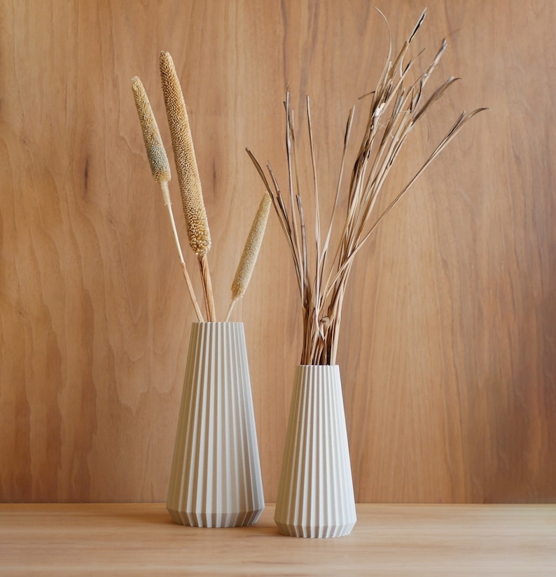 Deux vases en céramique blanche avec un design plissé, l'un contenant des tiges de blé séchées et l'autre des herbes séchées. Les vases sont sur une surface en bois.