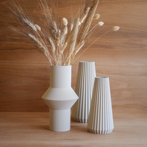 May include: Three white ceramic vases with a textured, ribbed design. The vases are arranged in a row on a wooden surface. A bouquet of dried grasses and wheat is in the tallest vase.