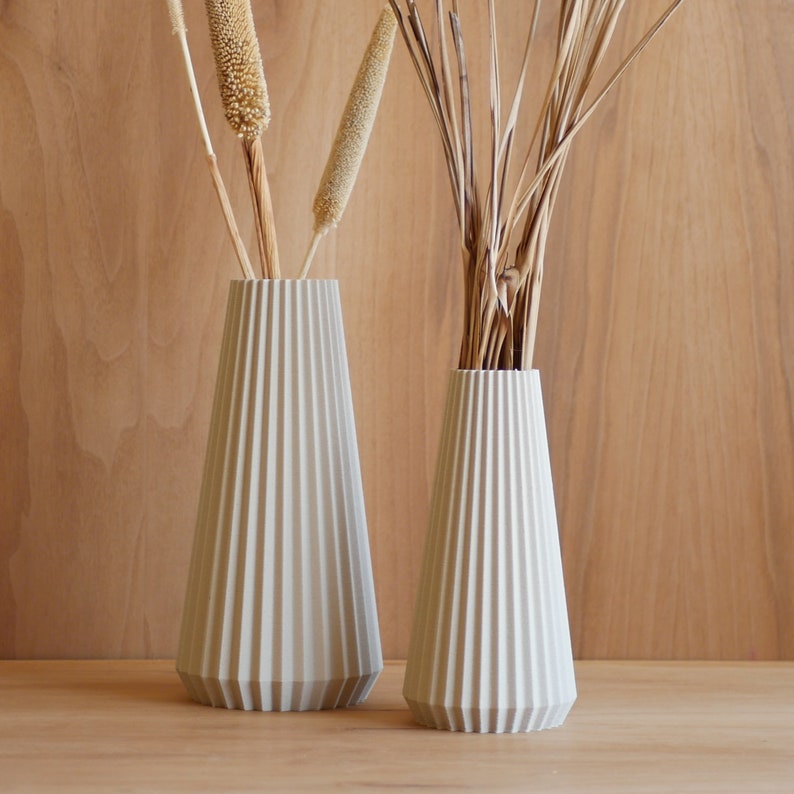 May include: Two white ceramic vases with a pleated design. The vases are empty and are sitting on a wooden surface.