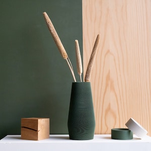 Puede incluir: Un jarrón verde oscuro con textura ondulada contiene tallos de flores secas beige sobre un fondo verde y madera.