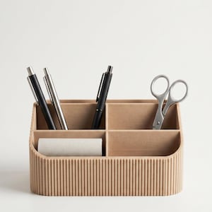 Puede incluir: Un organizador de escritorio beige con exterior acanalado y cuatro compartimentos. El organizador contiene dos bolígrafos negros, un bolígrafo plateado, unas tijeras plateadas y un pequeño rollo cilíndrico de papel o cinta adhesiva.